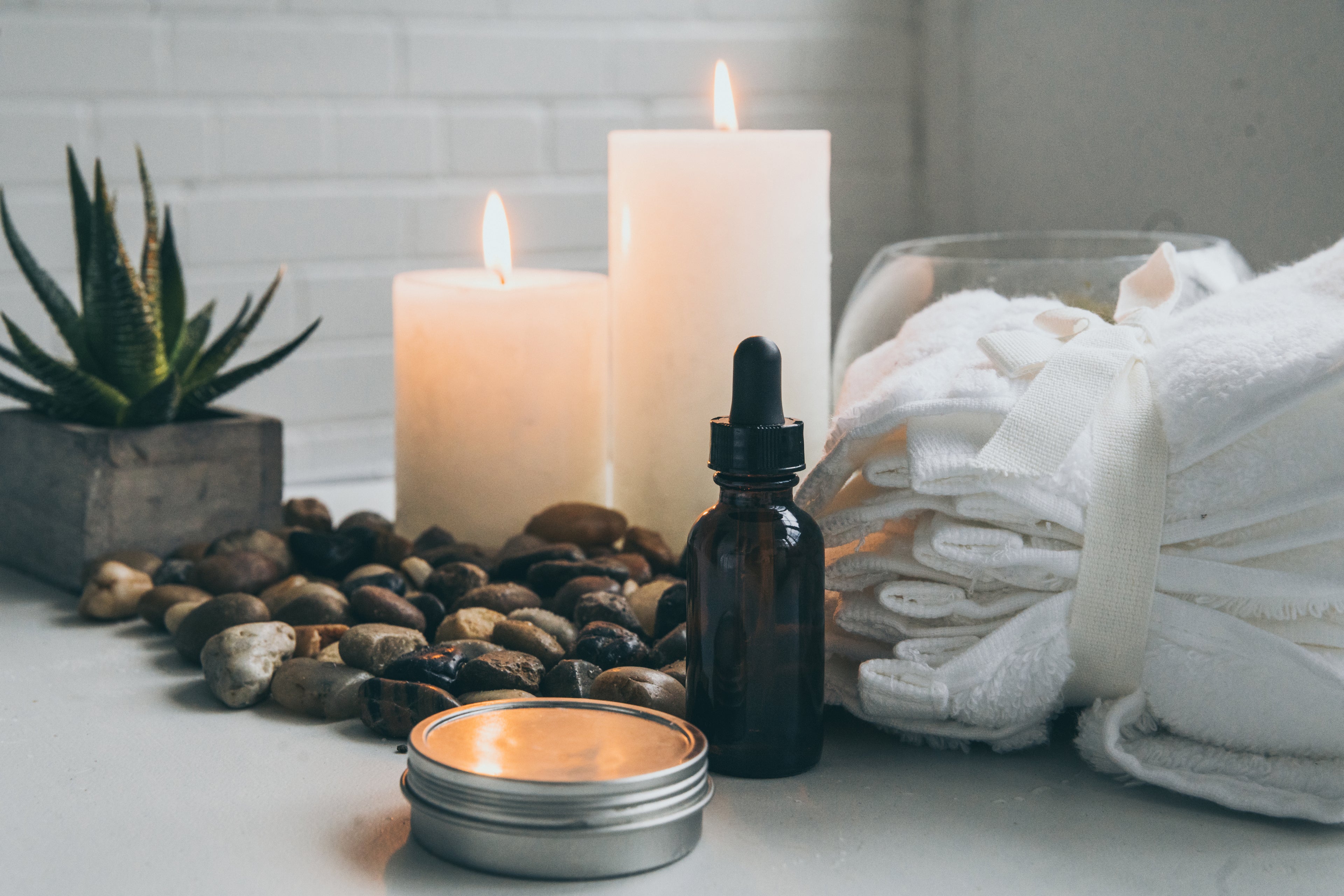 A serene spa setting with two lit candles, a small dropper bottle, folded white towels, and a round tin on a table. Stones and a small potted succulent are arranged nearby, creating a tranquil atmosphere.